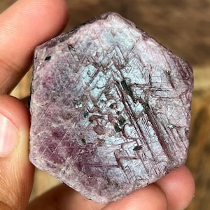 Peut inclure: Une pierre précieuse de rubis brute, de forme hexagonale et de couleur rose foncé. La surface présente des motifs linéaires complexes et de petites inclusions noires, créant un aspect texturé. La pierre est tenue dans une main.