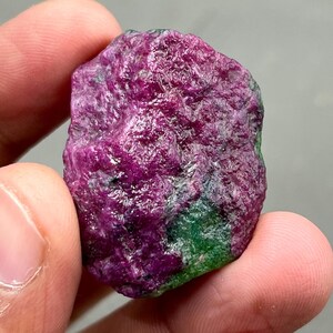 Może przedstawiać: Surowy kamień szlachetny rubinu w głębokim odcieniu magenty z zielonymi akcentami. Surowy minerał trzymany między palcami, ukazujący naturalną, nieoszlifowaną fakturę. Unikalny dodatek do każdej kolekcji kamieni.