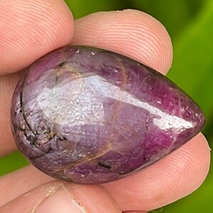 Può includere: Una pietra preziosa di rubino lucidata a forma di lacrima, di colore rosso intenso e con inclusiones nere. La pietra preziosa è traslucida e ha una finitura lucida.