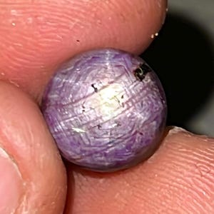 May include: A close-up of a purple star ruby gemstone with a star-like pattern. The gemstone is round and has a smooth, polished surface.