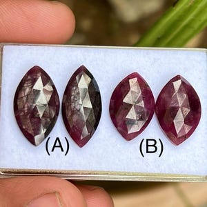Pode incluir: Quatro pedras preciosas em forma de marquesa com um tom vermelho profundo são exibidas em uma bandeja branca. Duas são rotuladas (A) e duas (B). As pedras têm superfícies facetadas, que capturam a luz e criam um efeito brilhante. As pedras são provavelmente para fazer joias.