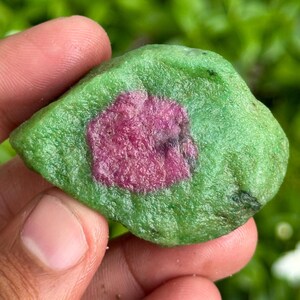以下が含まれることがあります： 鮮やかな緑色の外観と、中央に深いマゼンタ色の不規則なパッチがある、未加工の宝石。石の表面は質感があり、色と形の自然なバリエーションを示しています。コレクター向けのユニークな作品です。