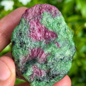 May include: A natural gemstone with a striking colour combination. The stone displays a blend of deep red and green shades, with black markings. The surface has a textured look, highlighting the mineral's natural formation. The gemstone is held in a hand.