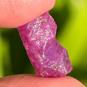 Op de afbeelding: Een ruwe, ongeslepen robijn edelsteen. De robijn heeft een levendige roodroze kleur, met een ruw, getextureerd oppervlak. De edelsteen wordt tussen twee vingers gehouden tegen een vage groene achtergrond.