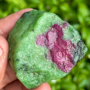 Peut inclure: Une pierre précieuse brute avec une matrice verte vibrante et une inclusion rouge profond proéminente. La pierre a un aspect naturel et non poli, mettant en valeur sa beauté brute. Le fond est vert flou.