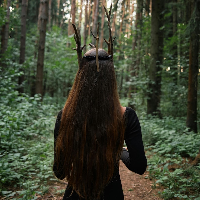 May include: A person with long brown hair wearing a black dress and a crown made of twigs, walking on a path in a forest. The crown has several upright twig elements.
