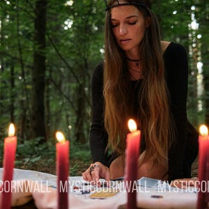 Puede incluir: Un conjunto de siete velas rojas cil&iacute;ndricas y altas dispuestas en semic&iacute;rculo sobre un pa&ntilde;o blanco, cada una con una llama encendida. El texto MYSTICORNWALL est&aacute; impreso en blanco en la parte inferior de la imagen, con una persona de negro.