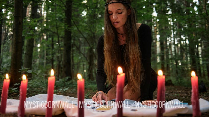 Puede incluir: Un conjunto de siete velas rojas cil&iacute;ndricas y altas dispuestas en semic&iacute;rculo sobre un pa&ntilde;o blanco, cada una con una llama encendida. El texto MYSTICORNWALL est&aacute; impreso en blanco en la parte inferior de la imagen, con una persona de negro.