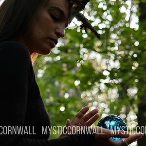 Puede incluir: Una esfera de cristal azul transl&uacute;cido y lisa sostenida en las palmas de las manos. La esfera refleja la luz y tiene una forma redondeada. El texto MYSTICCORNWALL est&aacute; impreso en letras may&uacute;sculas blancas en la parte inferior de la imagen.