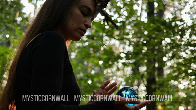 Puede incluir: Una esfera de cristal azul transl&uacute;cido y lisa sostenida en las palmas de las manos. La esfera refleja la luz y tiene una forma redondeada. El texto MYSTICCORNWALL est&aacute; impreso en letras may&uacute;sculas blancas en la parte inferior de la imagen.