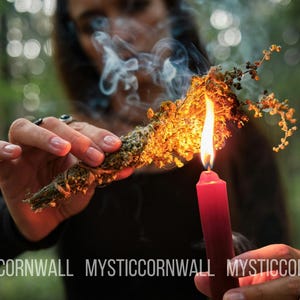 Puede incluir: Un manojo de hierbas verdes secas atadas con cuerda es sostenido y encendido por una vela roja, produciendo una llama naranja brillante y un rastro de humo blanco. El texto MYSTIC CORNWALL est&aacute; impreso en blanco en la parte inferior.