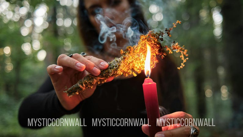 Puede incluir: Un manojo de hierbas verdes secas atadas con cuerda es sostenido y encendido por una vela roja, produciendo una llama naranja brillante y un rastro de humo blanco. El texto MYSTIC CORNWALL est&aacute; impreso en blanco en la parte inferior.
