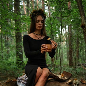 May include: A woman in a black dress and floral crown sits on a log in a forest. She holds a wooden bowl and a small piece of mushroom. The scene is set amongst green trees, with a natural, earthy aesthetic.