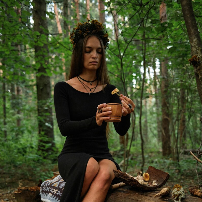 May include: A woman in a black dress and floral crown sits on a log in a forest. She holds a wooden bowl and a small piece of mushroom. The scene is set amongst green trees, with a natural, earthy aesthetic.