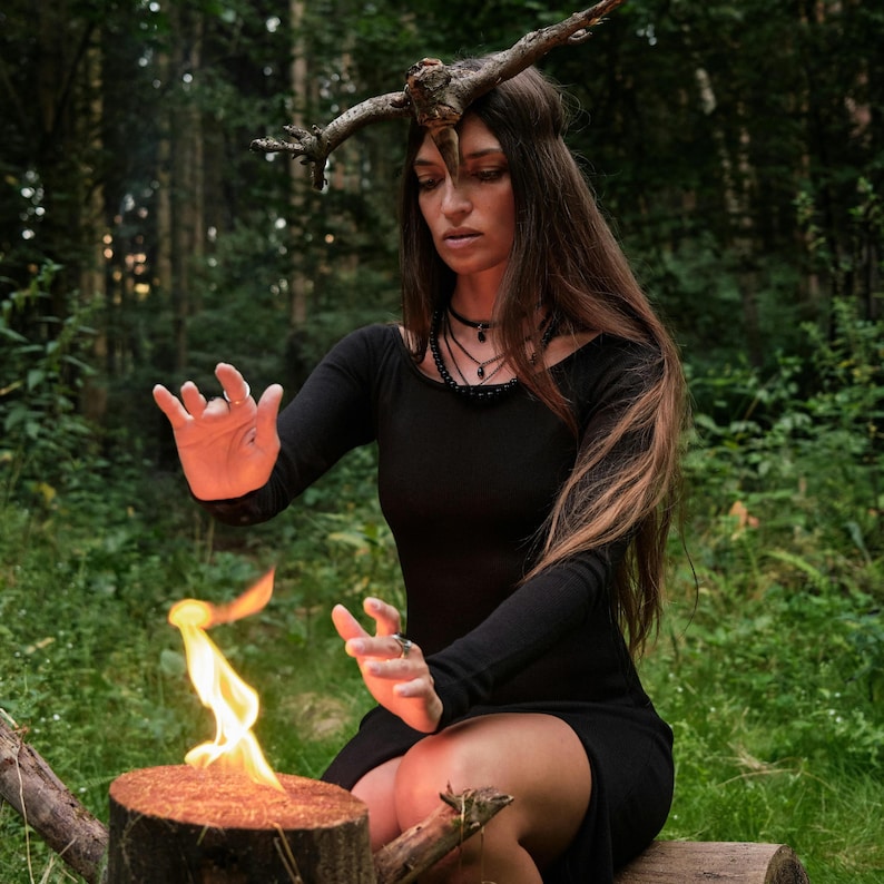May include: A woman in a black dress and a twig headpiece is positioned in a forest setting. A small fire burns on a wooden stump in front of her. She is wearing a black necklace and has long brown hair.
