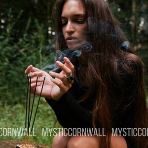 Puede incluir: Cinco varitas de incienso negras y finas se mantienen erguidas en un soporte de madera, emitiendo volutas de humo gris. El texto MYSTICCORNWALL est&aacute; impreso en blanco en la parte inferior. Una persona de pelo largo casta&ntilde;o viste una prenda negra.