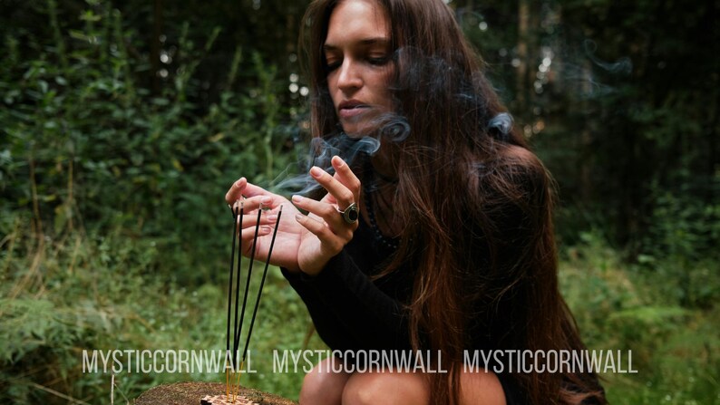 Puede incluir: Cinco varitas de incienso negras y finas se mantienen erguidas en un soporte de madera, emitiendo volutas de humo gris. El texto MYSTICCORNWALL est&aacute; impreso en blanco en la parte inferior. Una persona de pelo largo casta&ntilde;o viste una prenda negra.