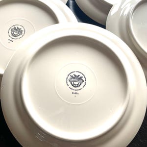 May include: A collection of white ceramic plates. The underside of each plate features a circular emblem with text and the word "India". The plates are arranged on a dark surface.