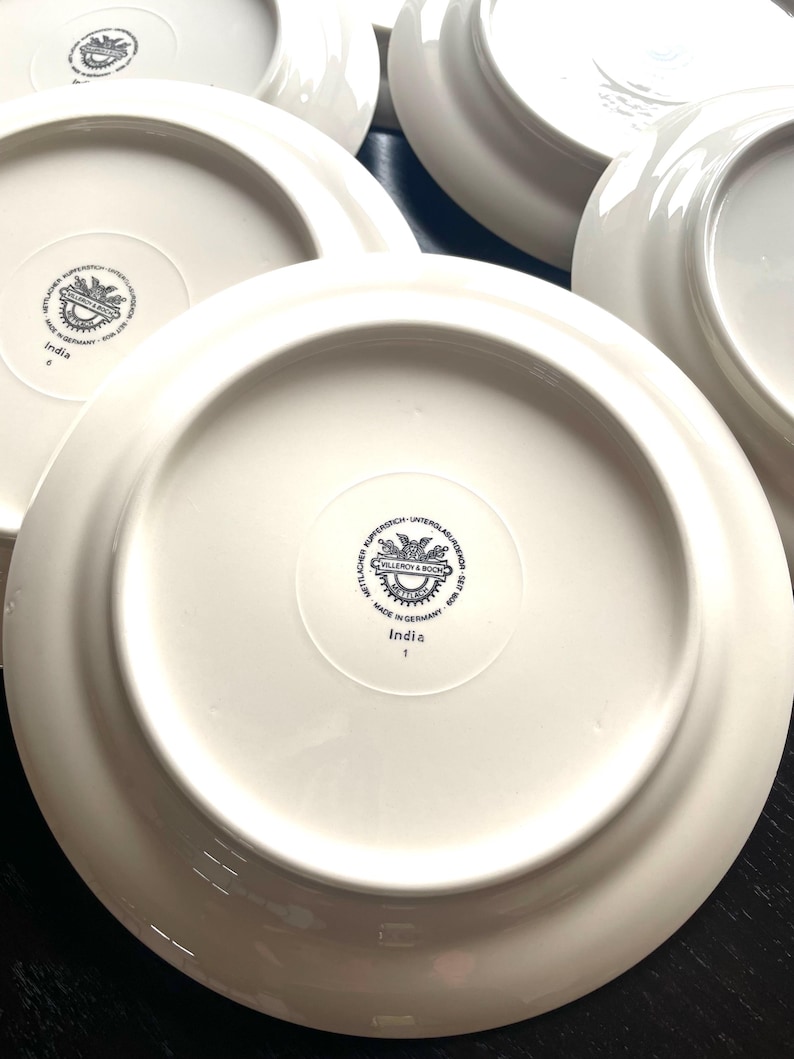 May include: A collection of white ceramic plates. The underside of each plate features a circular emblem with text and the word "India". The plates are arranged on a dark surface.