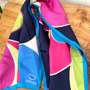 May include: A colorful silk scarf with a geometric design. The scarf features a vibrant mix of pink, blue, navy, white, and lime green. The word "OSCAR" is printed on a blue section of the scarf.