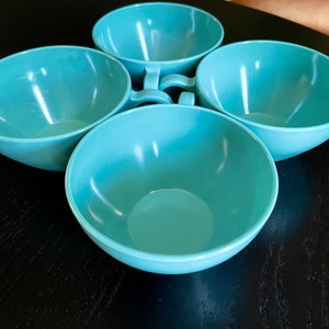 May include: Four turquoise-colored bowls with handles, arranged in a circular pattern. The bowls have a smooth, glossy finish and are empty. The handles are small and curved, and the bowls are sitting on a dark surface.