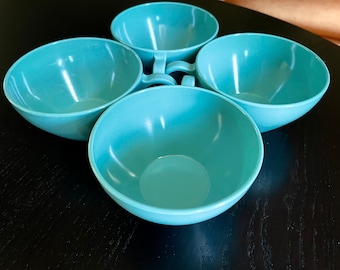 May include: Four turquoise-colored bowls with handles, arranged in a circular pattern. The bowls have a smooth, glossy finish and are empty. The handles are small and curved, and the bowls are sitting on a dark surface.