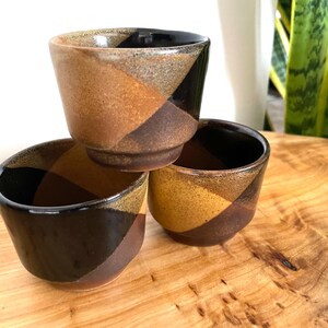 May include: Three ceramic cups stacked together. Each cup has a unique color scheme, with sections of brown, tan, and black. The cups are on a wooden surface, with a white background and greenery in the background.