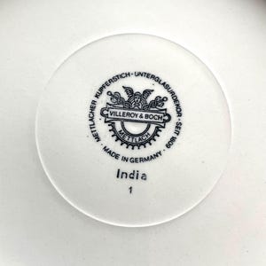 May include: Close-up of a white ceramic plate with a central circular emblem. The emblem features the text "Villeroy & Boch" and "Made in Germany." Below the emblem, the word "India" and the number "1" are printed. The plate has a simple, clean design.