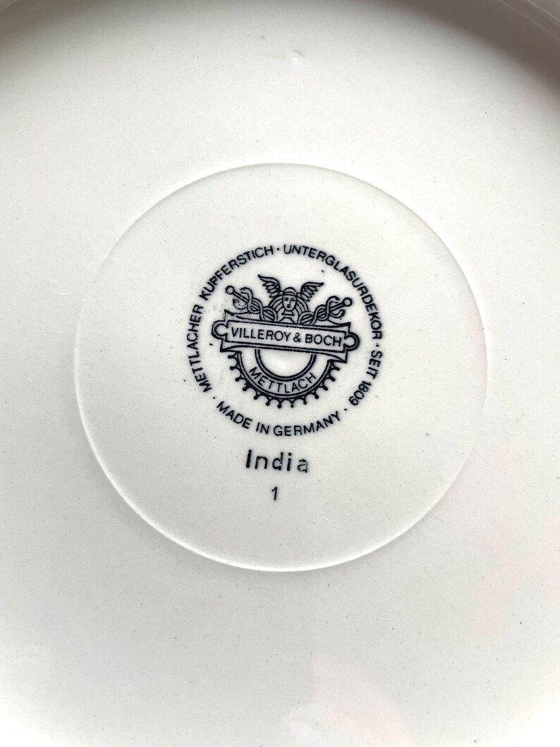 May include: Close-up of a white ceramic plate with a central circular emblem. The emblem features the text "Villeroy & Boch" and "Made in Germany." Below the emblem, the word "India" and the number "1" are printed. The plate has a simple, clean design.