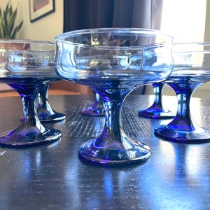 May include: Set of four cobalt blue coupe glasses. These vintage-style glasses have a wide, shallow bowl and a stemmed base. The glasses are made of clear glass with a deep blue hue, perfect for serving cocktails or champagne.