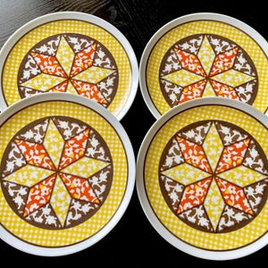 May include: Four round plates with a white rim, featuring a yellow gingham border and a central starburst design. The starburst is composed of brown, orange, yellow, and white floral patterns. A vintage, decorative tableware set.