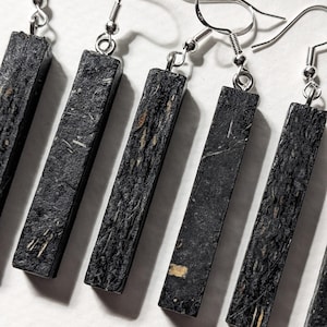 May include: A set of rectangular black earrings with silver-colored fish hook ear wires. The earrings have a textured surface with flecks of tan and white. The earrings are displayed on a white surface.