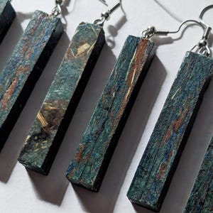 Könnte beinhalten: Nahaufnahme mehrerer rechteckiger Ohrringe mit silbernen Haken. Die Ohrringe bestehen aus einem dunkelblauen Material mit Streifen in Blau, Braun und Beige. Die Ohrringe sind diagonal vor einem weißen Hintergrund angeordnet.