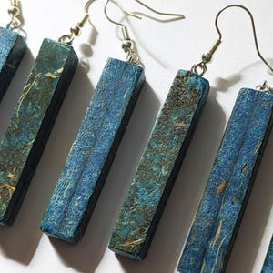 Puede incluir: Un conjunto de pendientes rectangulares con un acabado texturizado azul y turquesa. Los pendientes están suspendidos de ganchos para la oreja de color plateado. Los pendientes tienen un aspecto rústico y natural.