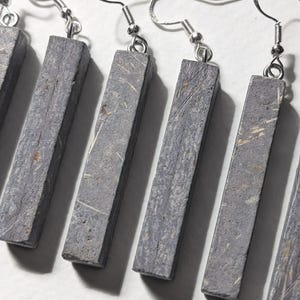 May include: A set of rectangular gray earrings with silver-colored fish hook ear wires. The earrings have a textured surface with light-colored streaks. The earrings are displayed on a white surface.
