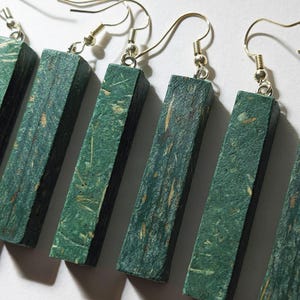 May include: Close-up of several green rectangular earrings with silver-colored hooks. The earrings have a textured surface with flecks of tan and brown. The earrings are displayed against a white background. The earrings are a unique accessory.