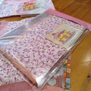 May include: A close-up of a pink and white fabric with a floral pattern. The fabric is folded and wrapped in clear plastic with a pink and white polka dot ribbon. A small card with a picture of a cake and text is visible.