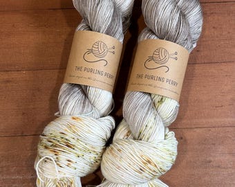 May include: Two skeins of yarn with brown paper labels. The yarn is a mix of white, gray, and tan colors. The labels read "THE PURLING PERRY" with a logo of a ball of yarn and knitting needles. The yarn is on a brown wooden surface.
