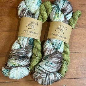 May include: Two skeins of yarn with a mix of colors, including white, brown, green, and teal. Each skein has a brown paper label that reads "THE PURLING PERRY" and "Hand-dyed in the Jemison mountains."