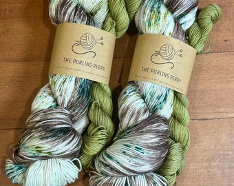 May include: Two skeins of yarn with a mix of colors, including white, brown, green, and teal. Each skein has a brown paper label that reads "THE PURLING PERRY" and "Hand-dyed in the Jemison mountains."