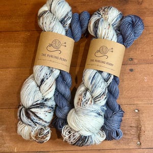 May include: Two skeins of yarn with a mix of white, brown, and blue fibers. Each skein has a dark blue section and a brown paper label that reads "THE PURLING PERRY". The yarn is displayed on a wooden surface.