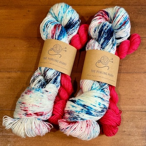 May include: Two skeins of hand-dyed yarn with a speckled pattern in white, blue, and red, with a bright pink accent. Each skein is wrapped with a brown paper label that reads "THE PURLING PERRY". The yarn is displayed on a wooden surface.