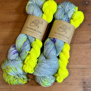 May include: Two skeins of hand-dyed yarn with a mix of colors. The yarn features a blend of gray, purple, and neon yellow. Each skein is wrapped with a brown paper label that reads "THE PURLING PERRY".