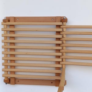 Puede incluir: Un tendedero plegable de madera de color claro montado en la pared. El tendedero está hecho de listones de madera horizontales y está fijado a una pared. El tendedero está parcialmente abierto, mostrando su diseño plegable. La madera tiene un acabado natural.