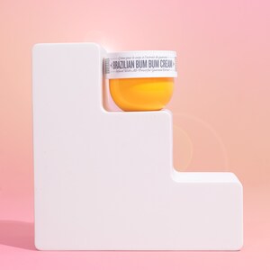 Op de afbeelding: Een gele pot Brazilian Bum Bum Cream op een wit platform. De pot heeft een wit etiket met de tekst "Creme pour le corps &agrave; l'extrait de guarana Brazilian Bum Bum Cream Infused With All-Powerful Guarana Extract".