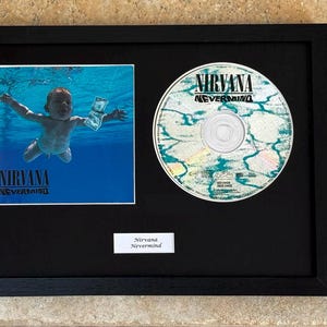 May include: A black framed display featuring the Nirvana Nevermind album cover art, a matching compact disc, and a small white plaque with the text Nirvana Nevermind printed in black. The album art depicts a baby swimming underwater toward a dollar bill.