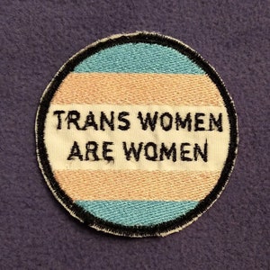 May include: A circular patch with the text "TRANS WOMEN ARE WOMEN" in black letters. The patch has a white background with a light blue and peach-colored stripe. The patch is outlined in black and sits on a purple surface.