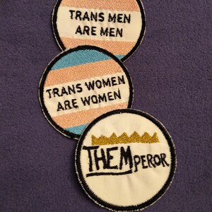 May include: Three round embroidered patches. The top patch is blue, pink, and white with the text "Trans Men Are Men". The middle patch is pink, blue, and white with the text "Trans Women Are Women". The bottom patch is white with a gold crown and the text "Them Emperor".