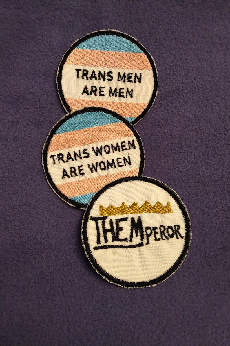 May include: Three round embroidered patches. The top patch is blue, pink, and white with the text "Trans Men Are Men". The middle patch is pink, blue, and white with the text "Trans Women Are Women". The bottom patch is white with a gold crown and the text "Them Emperor".