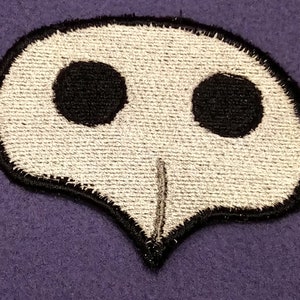 May include: A white embroidered patch with a black skull design. The skull has two black eyes and a small black nose.